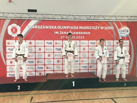Czterech młodych zawodników w białych judogach, z medalami na szyi stojacych na podium. W tle Ścianka z napisem Warszawska Olimpiada Młodziezy w Judo.